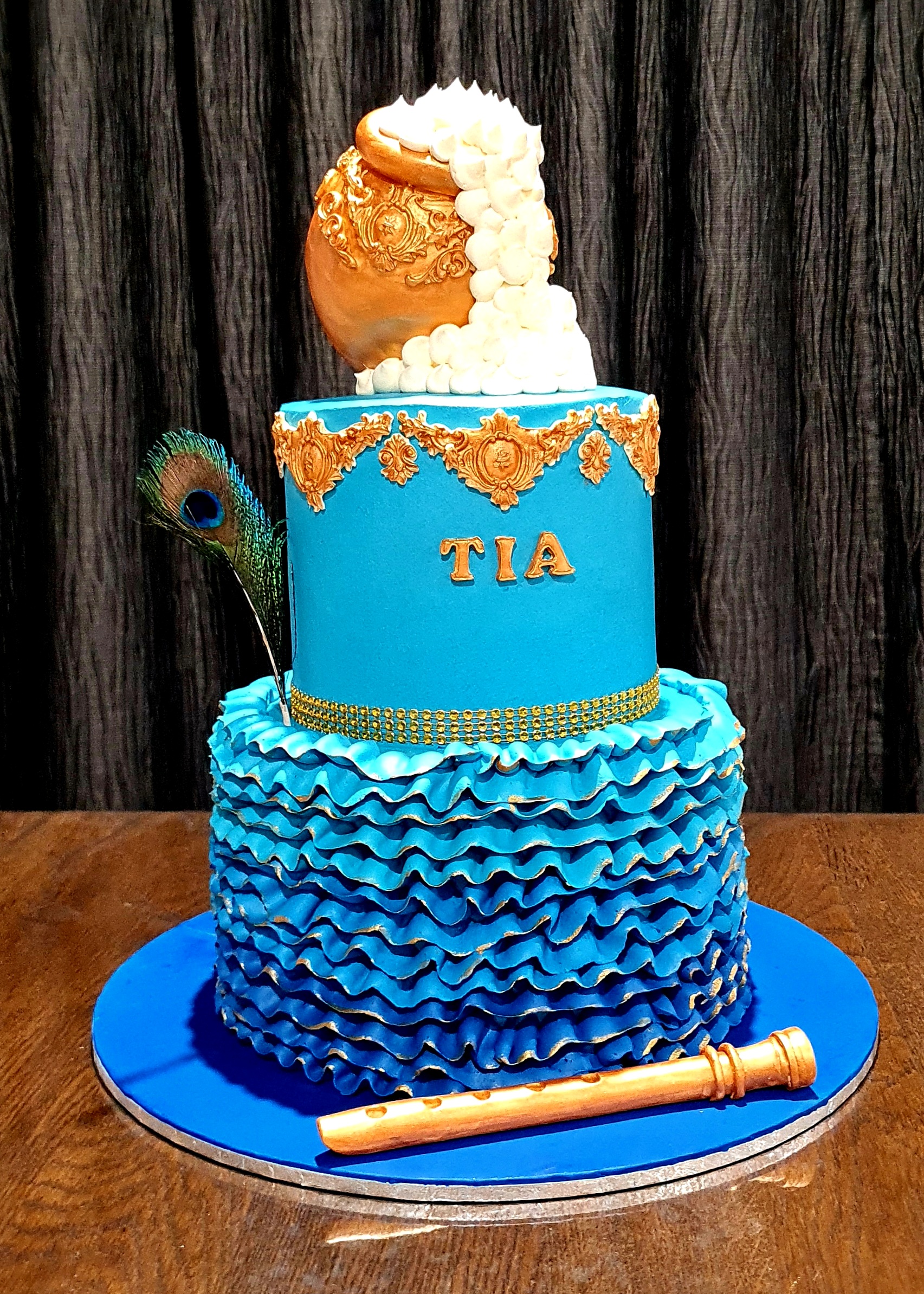 Krishna Theme Fusion Cake with blue tiers, gold Indian motifs, peacock feather and flute for Tia's celebration