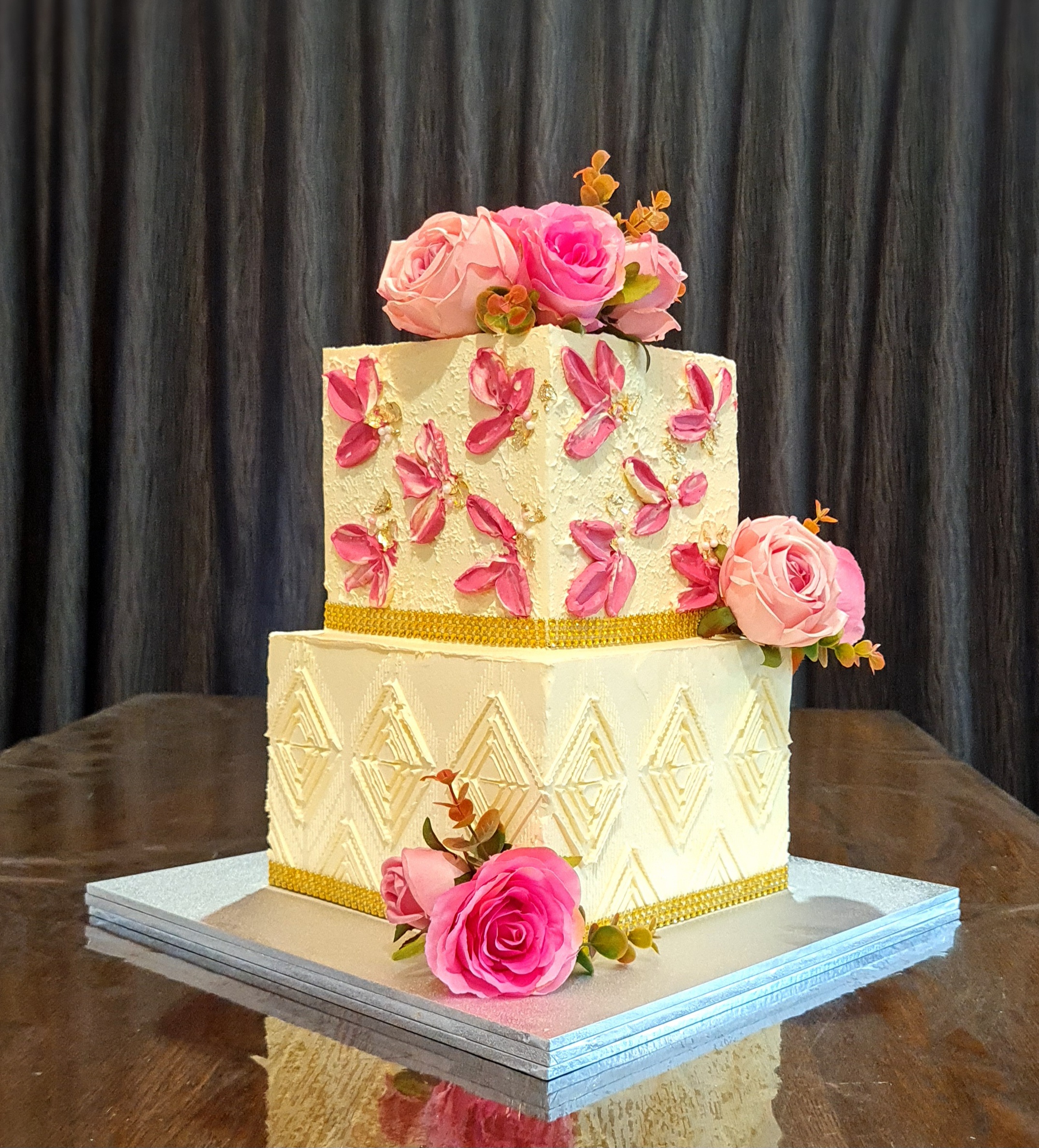 Two-tier square ivory cake with pink sugar flowers, butterflies, and gold ribbon for engagement/wedding